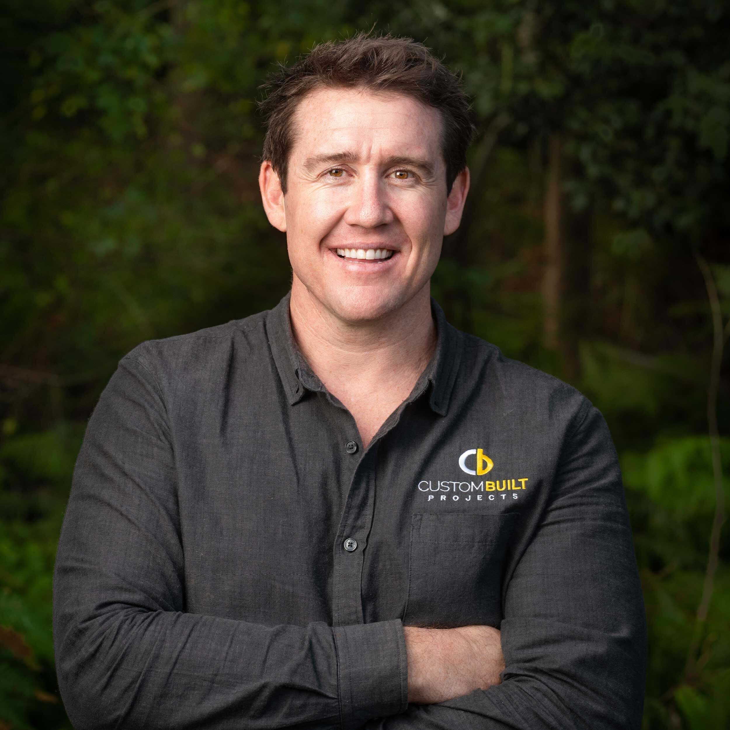 A smiling man in a gray shirt with a "Custom Built Projects" logo on it, standing outdoors with lush greenery blurred in the background. He appears confident and approachable near Port Stephens.