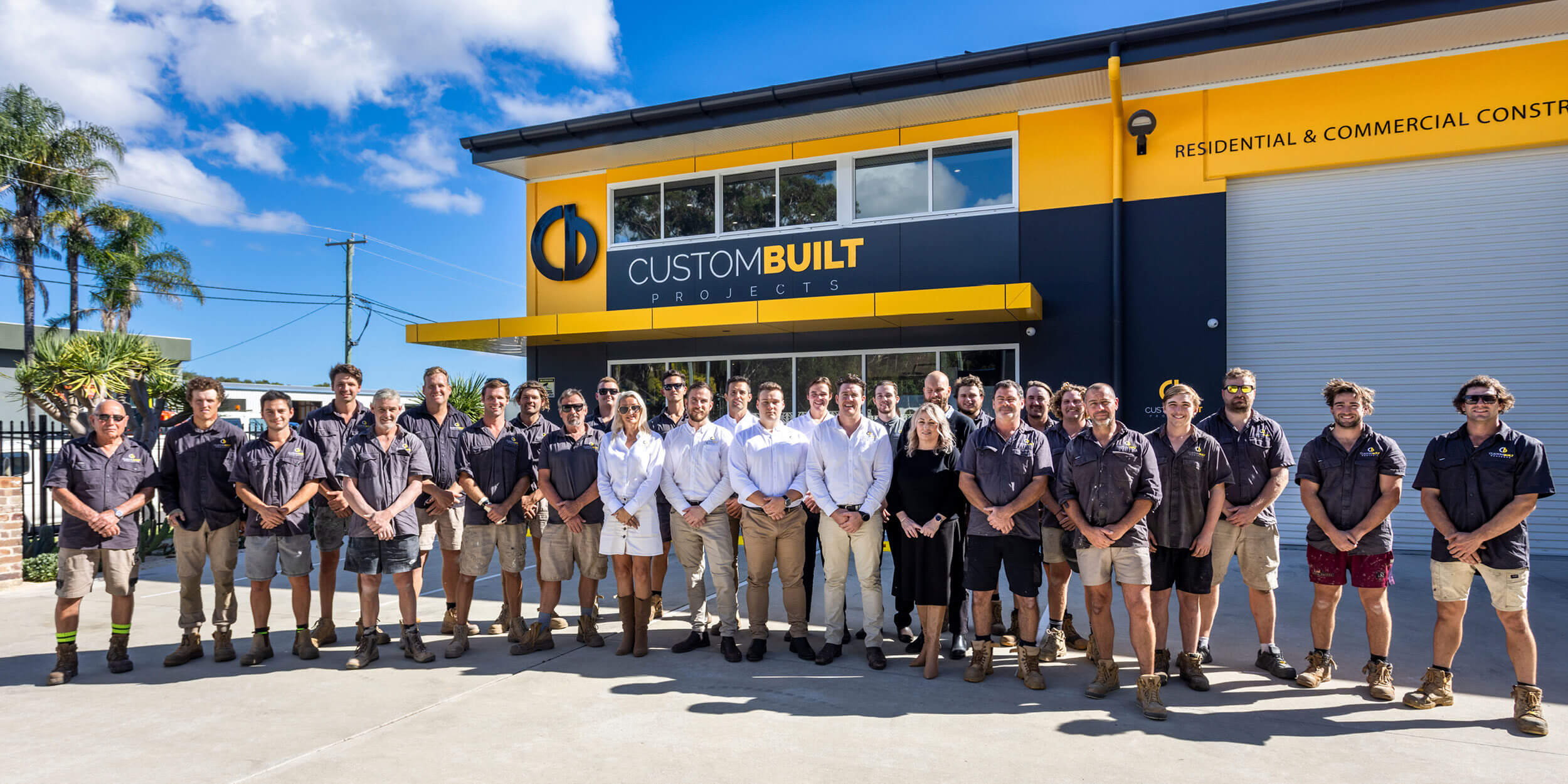 A group of approximately 20 construction workers in uniform, along with business professionals, stand in front of a building in Port Stephens with a sign reading "custom built projects," featuring industrial and office sections.