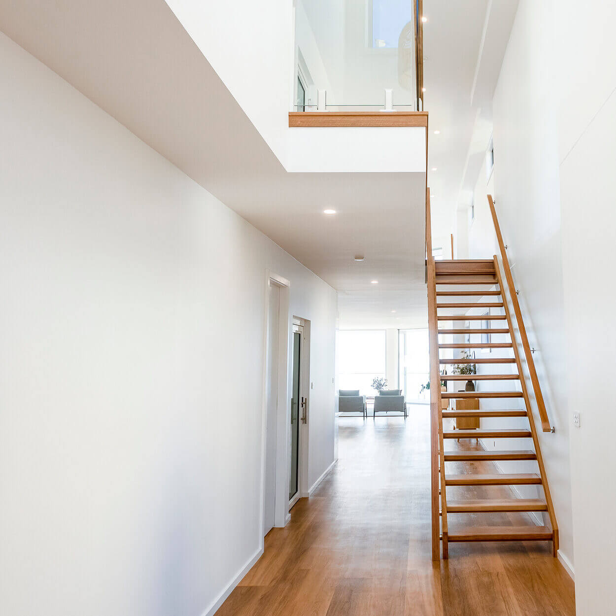 A modern, bright interior of a home featuring a custom-built wooden staircase leading to a mezzanine. The room has white walls, sleek hardwood floors, and is well-lit from large windows visible