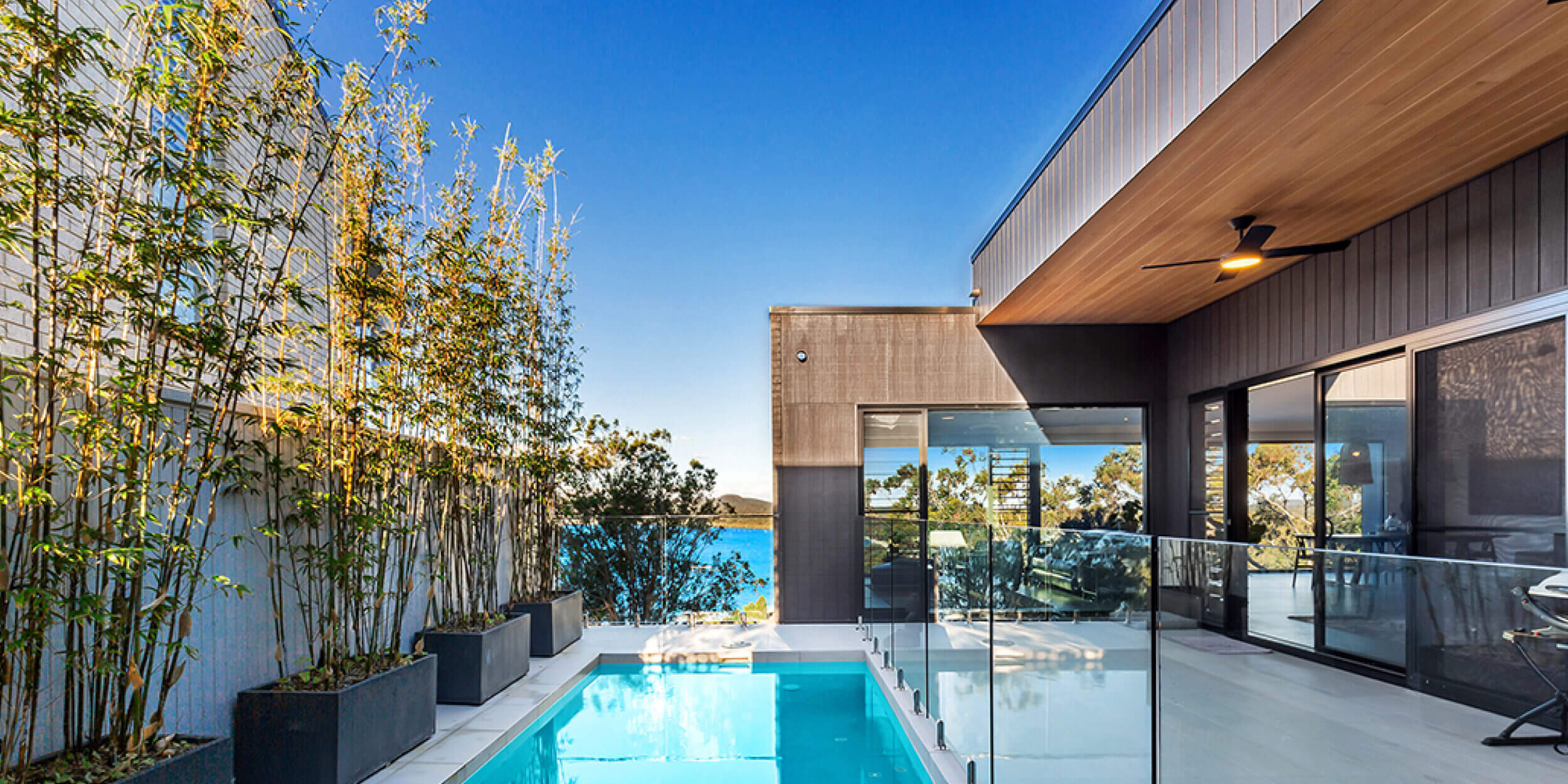 An outdoor area featuring a narrow swimming pool with clear blue water, surrounded by glass fencing. Adjacent is a modern house with dark wood panels and large sliding glass doors, located in Port Stephens. L