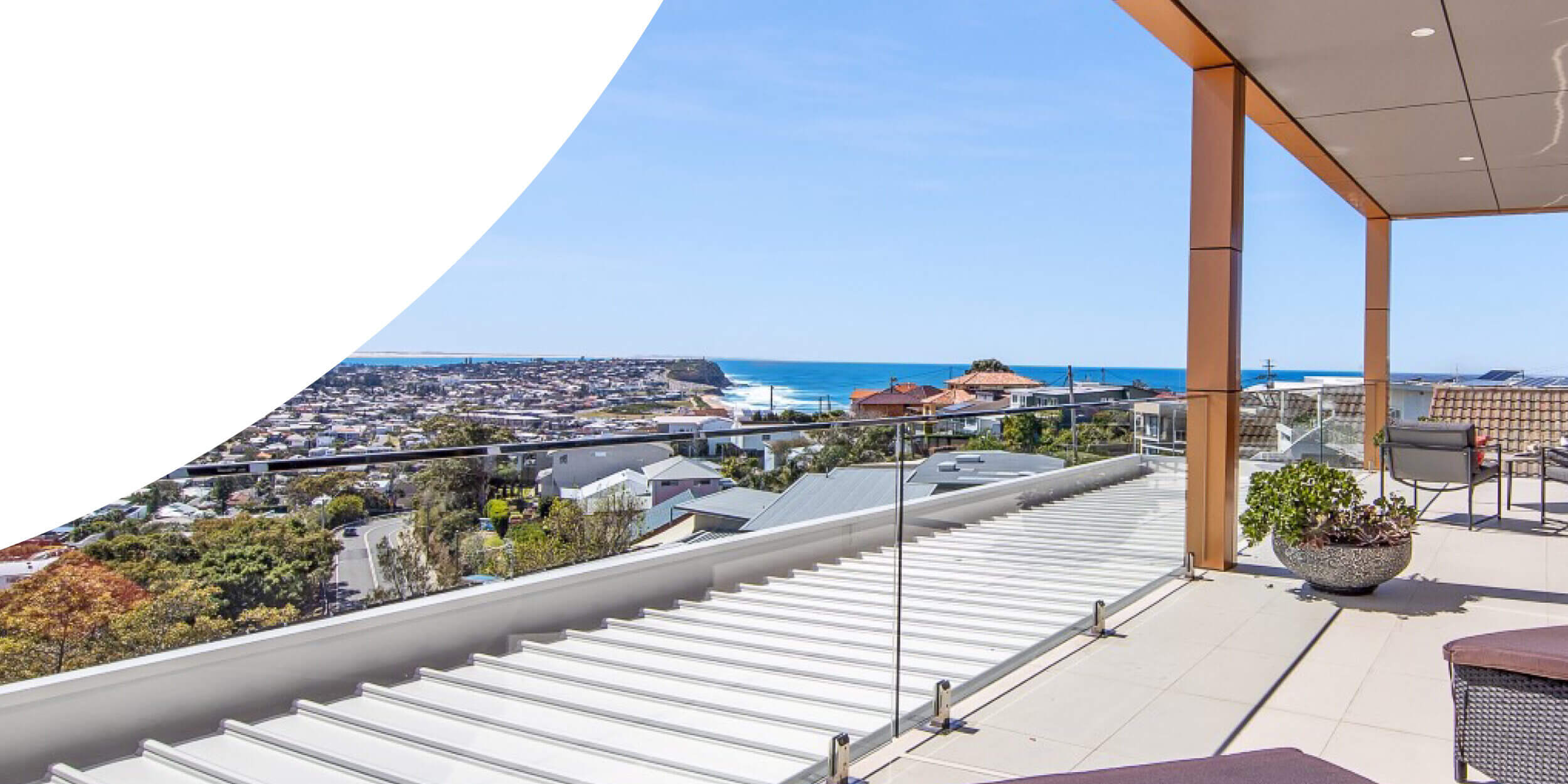 A spacious balcony overlooking the scenic coastal town of Port Stephens with clear skies, featuring modern outdoor furniture and glass railing, providing an unobstructed view of the blue ocean and surrounding houses.