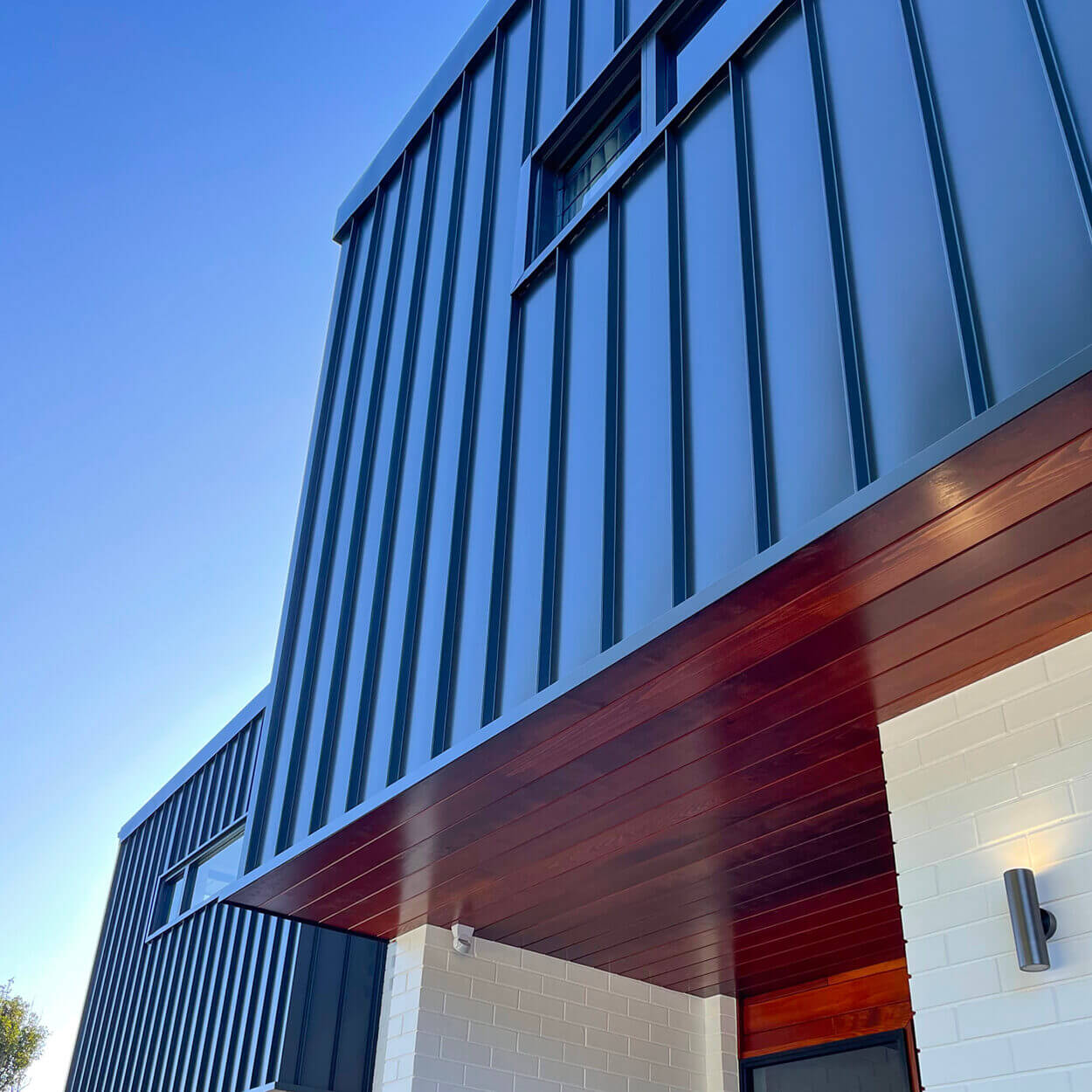 A modern architectural building corner view featuring dark blue vertical metal panels, a contrasting strip of warm-toned wood siding, and a section of white bricks, under a clear blue sky in Nelson Bay.