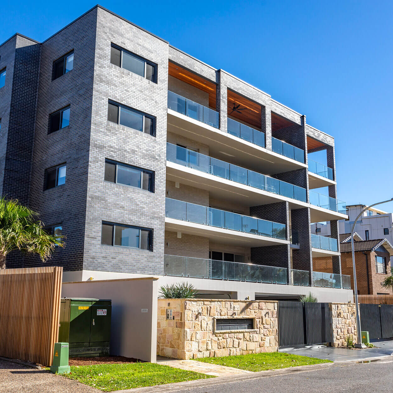 Modern multi-story residential building in Nelson Bay with a mix of stone and dark gray brick facade, featuring large balconies with glass railings and wood paneling under a clear blue sky.
