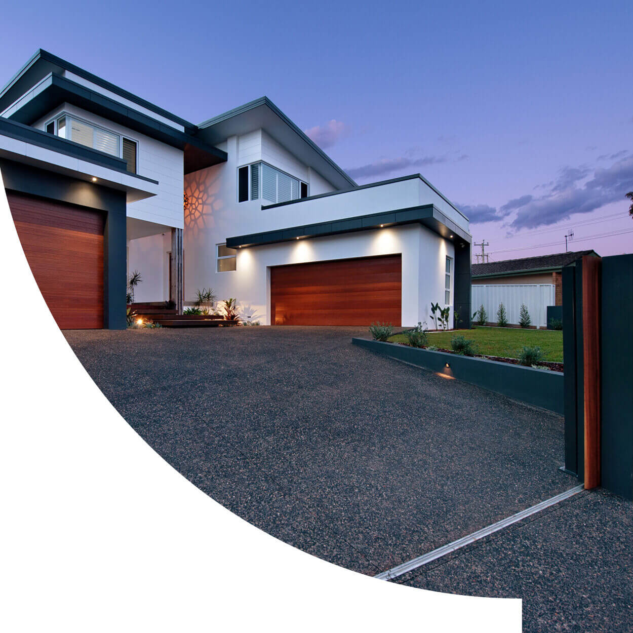 Modern two-story house in Nelson Bay at dusk, featuring a white exterior with wooden accents, a large garage door, and a well-lit driveway leading to a gate. The sky is vibrant with a
