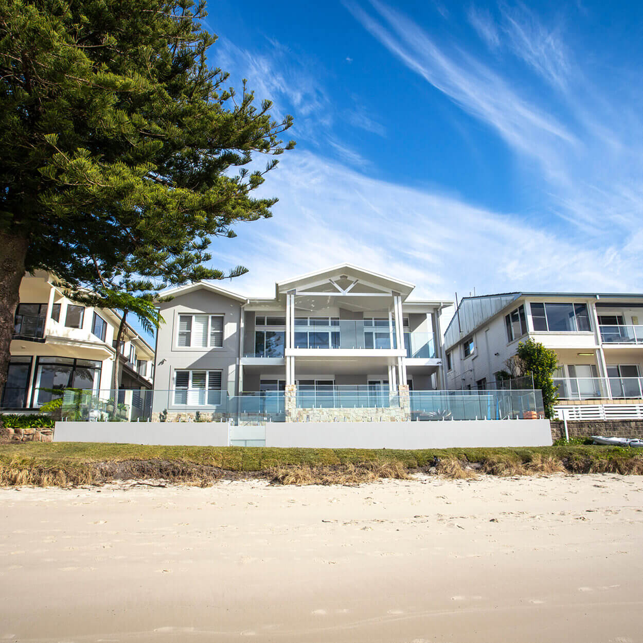 A luxurious beachfront house in Nelson Bay with large glass windows and a spacious balcony, flanked by other homes and a tall pine tree, under a clear blue sky streaked with wispy clouds.