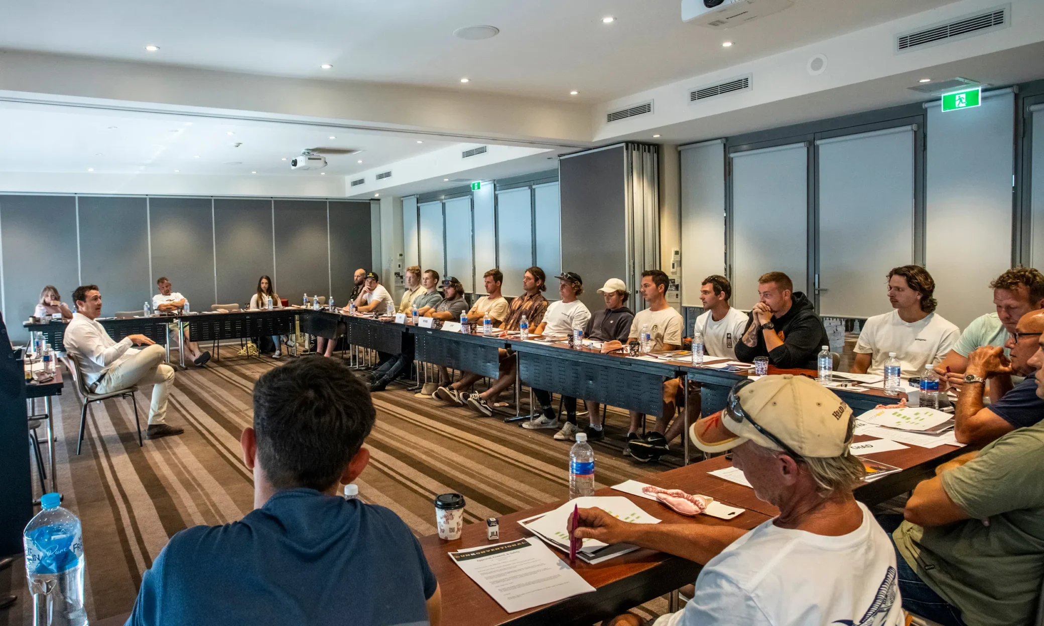 A professional meeting room with a group of people seated around a u-shaped table setup. They are attentively listening to a speaker seated at the head of the table, engaging in a discussion.