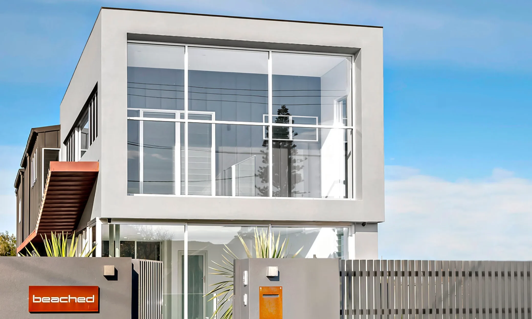 Modern two-story house in Newcastle with a prominent cubic upper floor featuring large windows. The aesthetic includes a minimalist white exterior and a small balcony with a terracotta planter. The logo "beached