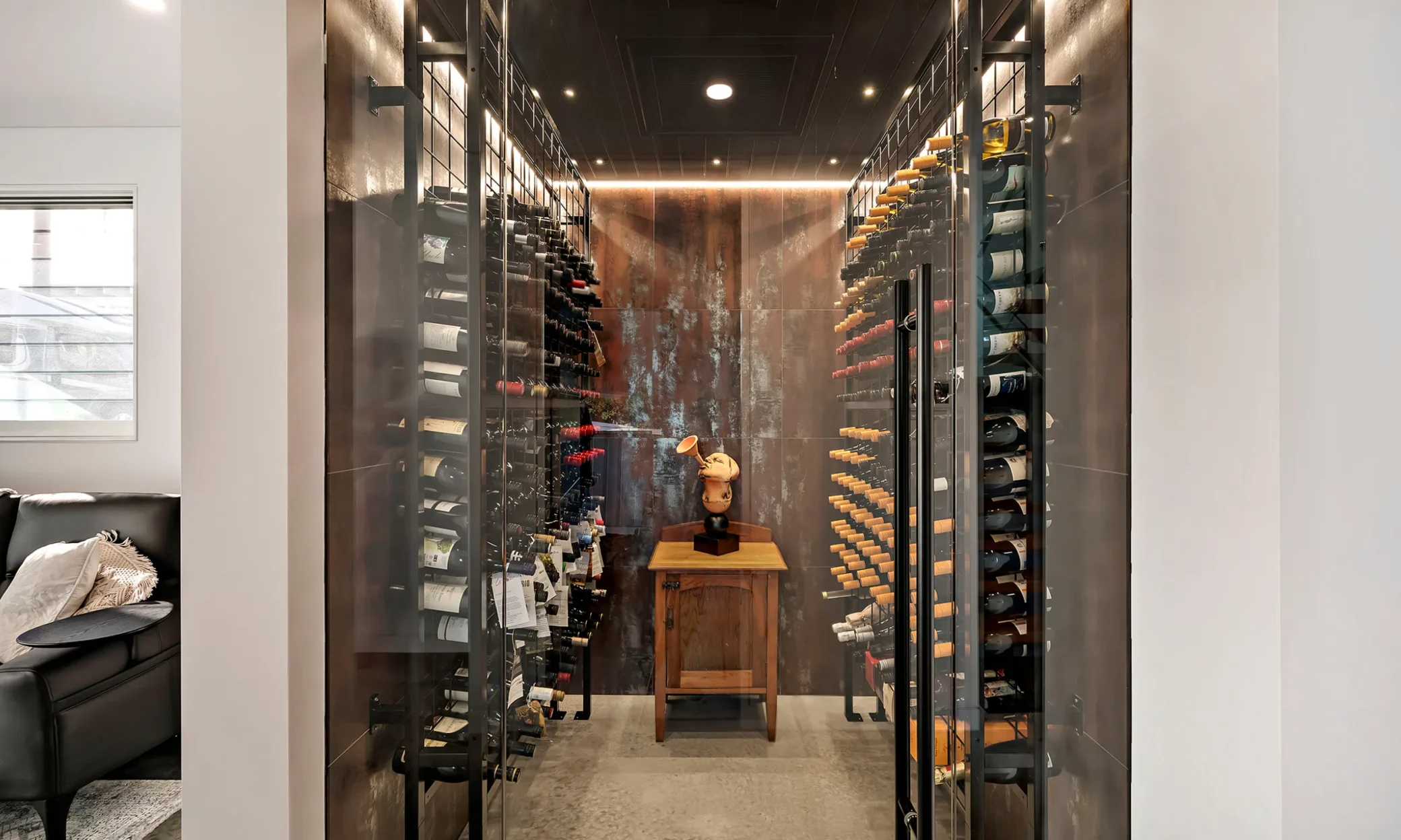 A modern wine cellar with floor-to-ceiling metal racks on each side filled with various wine bottles. A custom-built wood statue of a violinist centers the room with a spotlight overhead. The room features