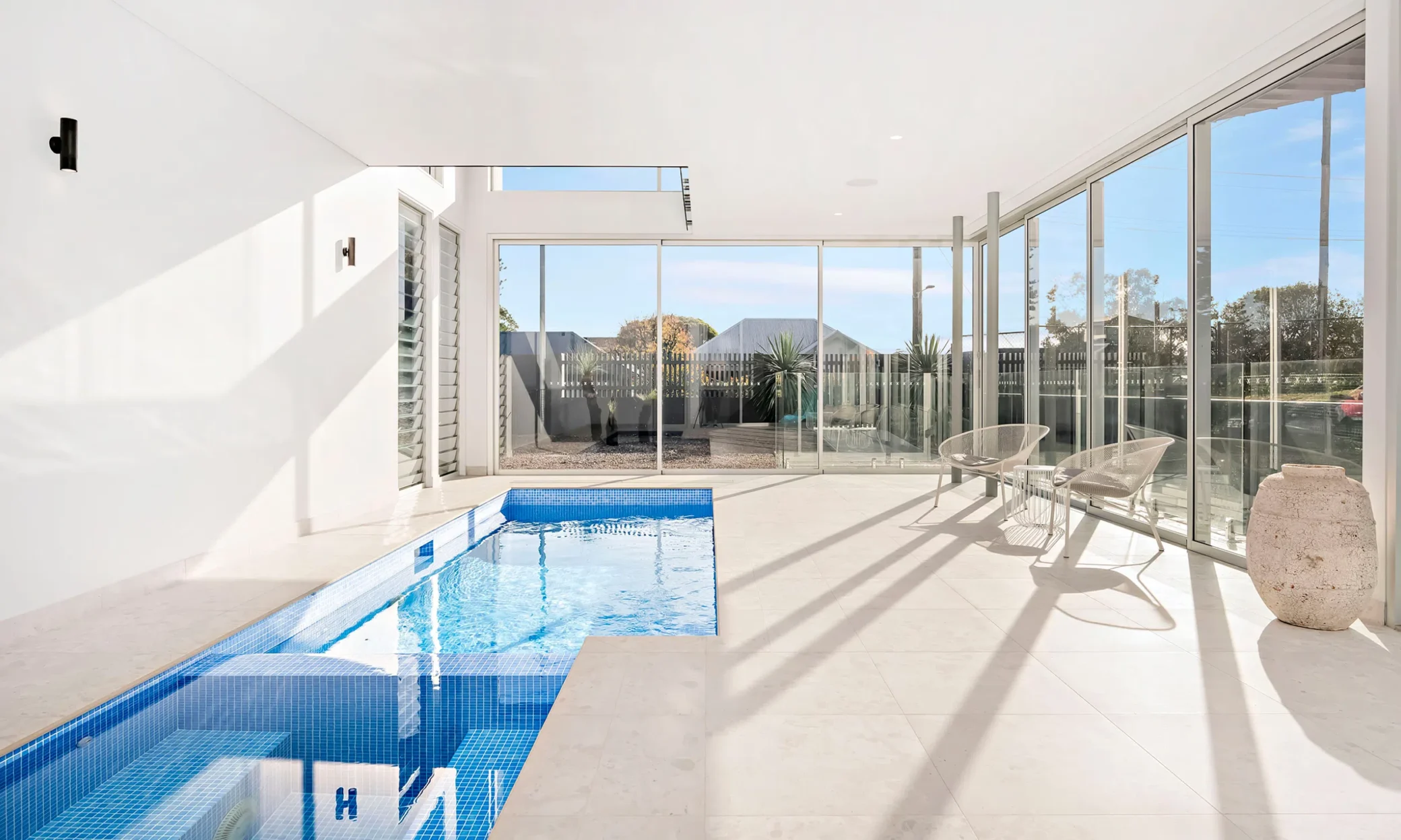 Modern indoor pool room in Newcastle with large glass sliding doors opening to an outdoor area. The room features a blue tiled pool, glossy white floor, two white chairs near a window, and bright natural light