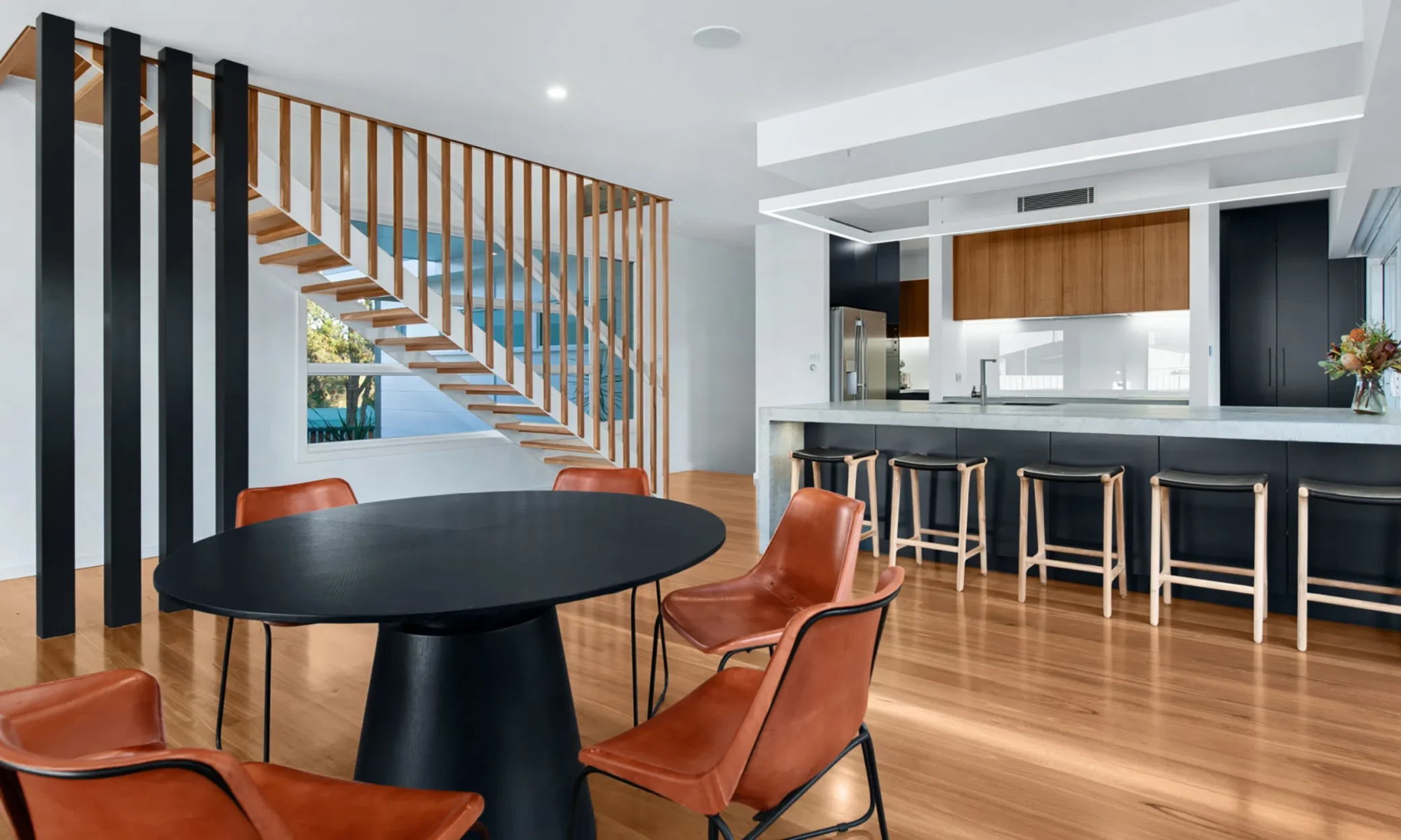 Modern kitchen and dining area featuring a black round table, brown leather chairs, a kitchen island with stools, and a wooden staircase with vertical slats in Newcastle. Bright, airy atmosphere enhanced by large windows