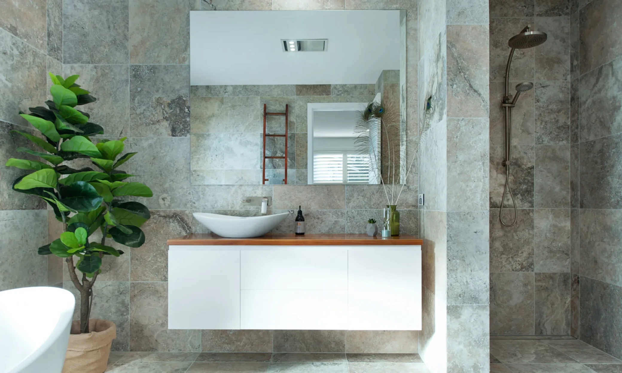 Modern bathroom with stone tile walls and floor, featuring a custom built white floating vanity with a vessel sink, a ladder towel rack, a large mirror, and a green potted plant next to a walk