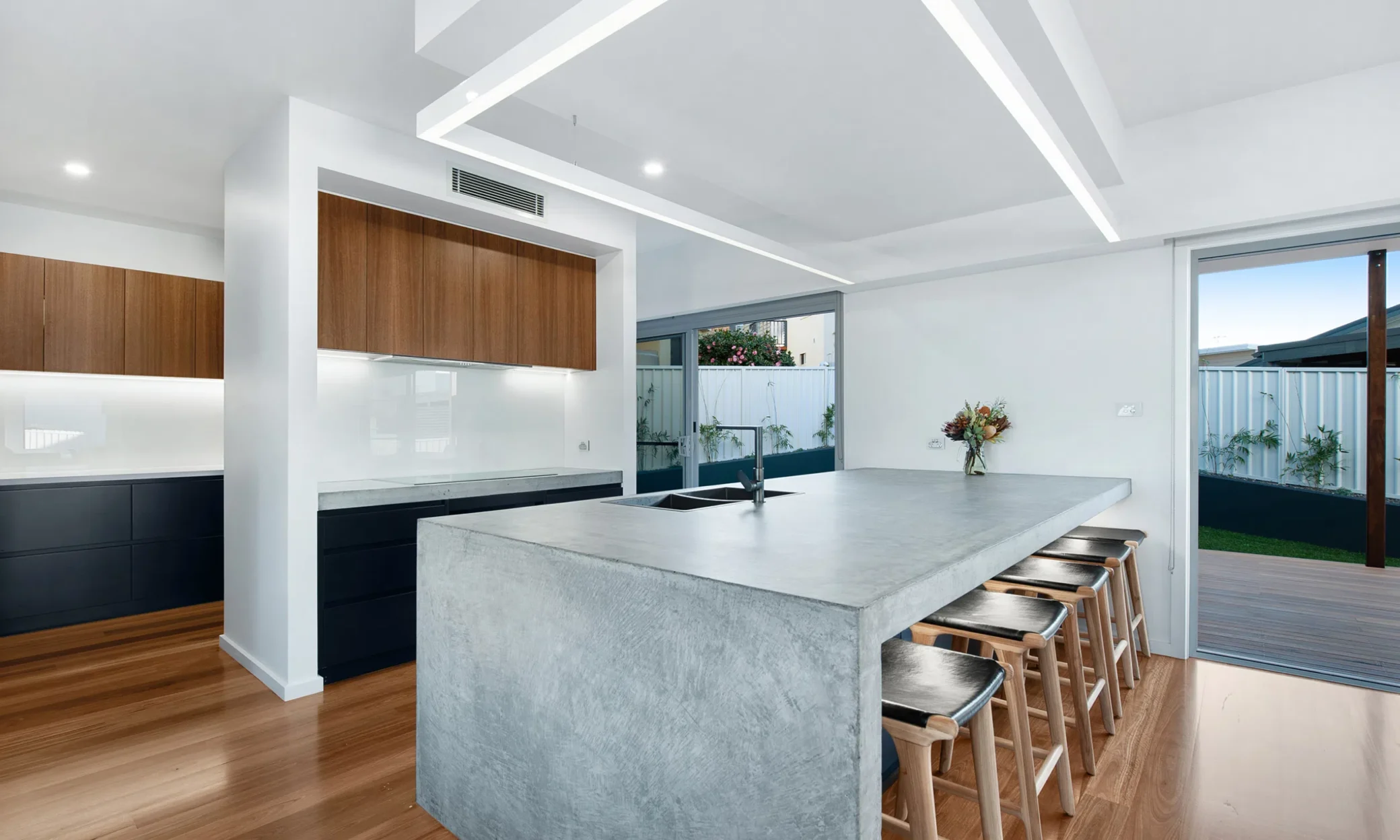 Modern kitchen interior featuring a large concrete island with bar stools, sleek wood cabinetry, and integrated appliances. The custom-built space opens to a scenic outdoor area through glass sliding doors, enhancing natural light.