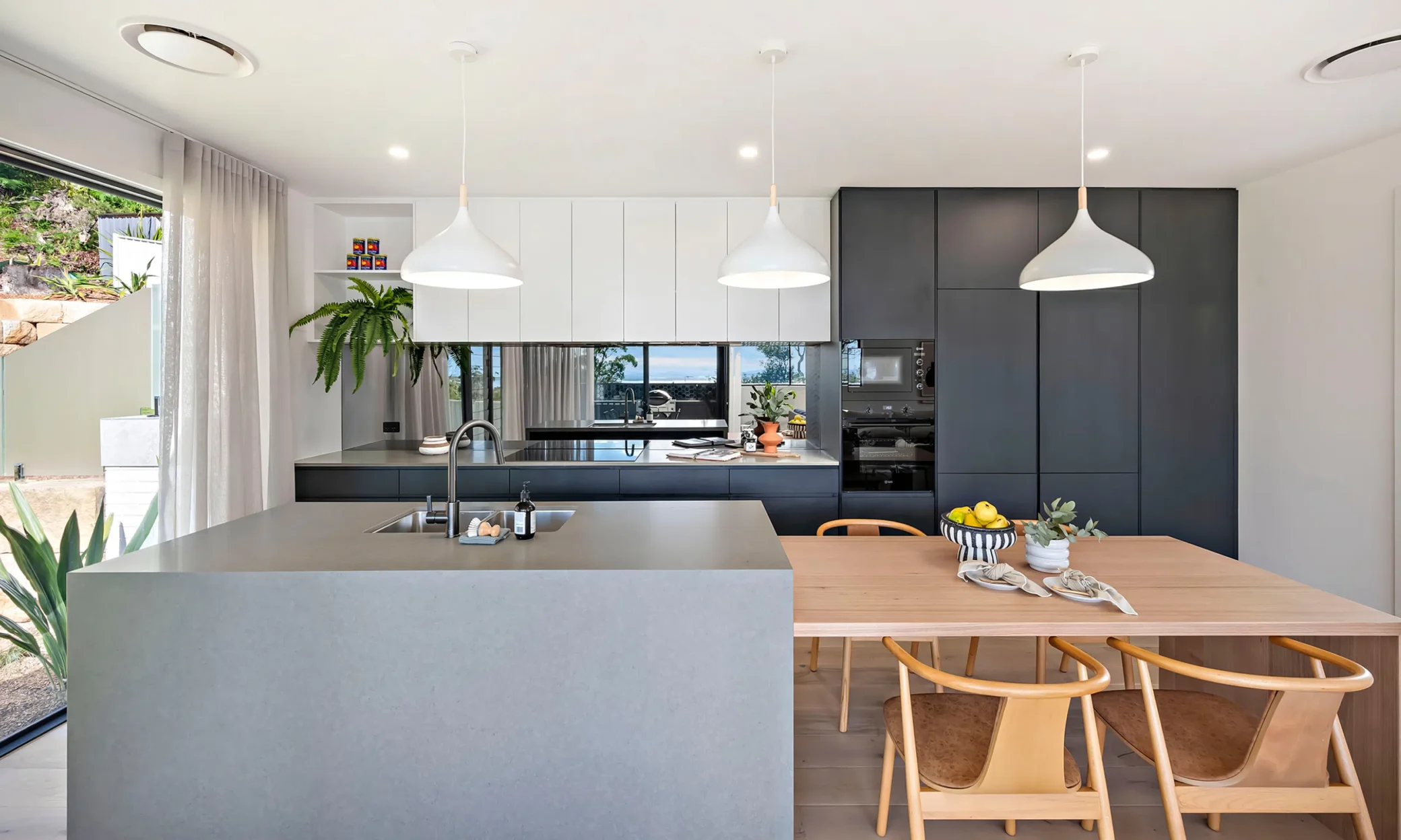 Modern kitchen with a central grey island, wooden dining table, and sleek black cabinets. White pendant lights hang above, with a view of greenery outside through large windows. Bright and airy ambiance, featuring