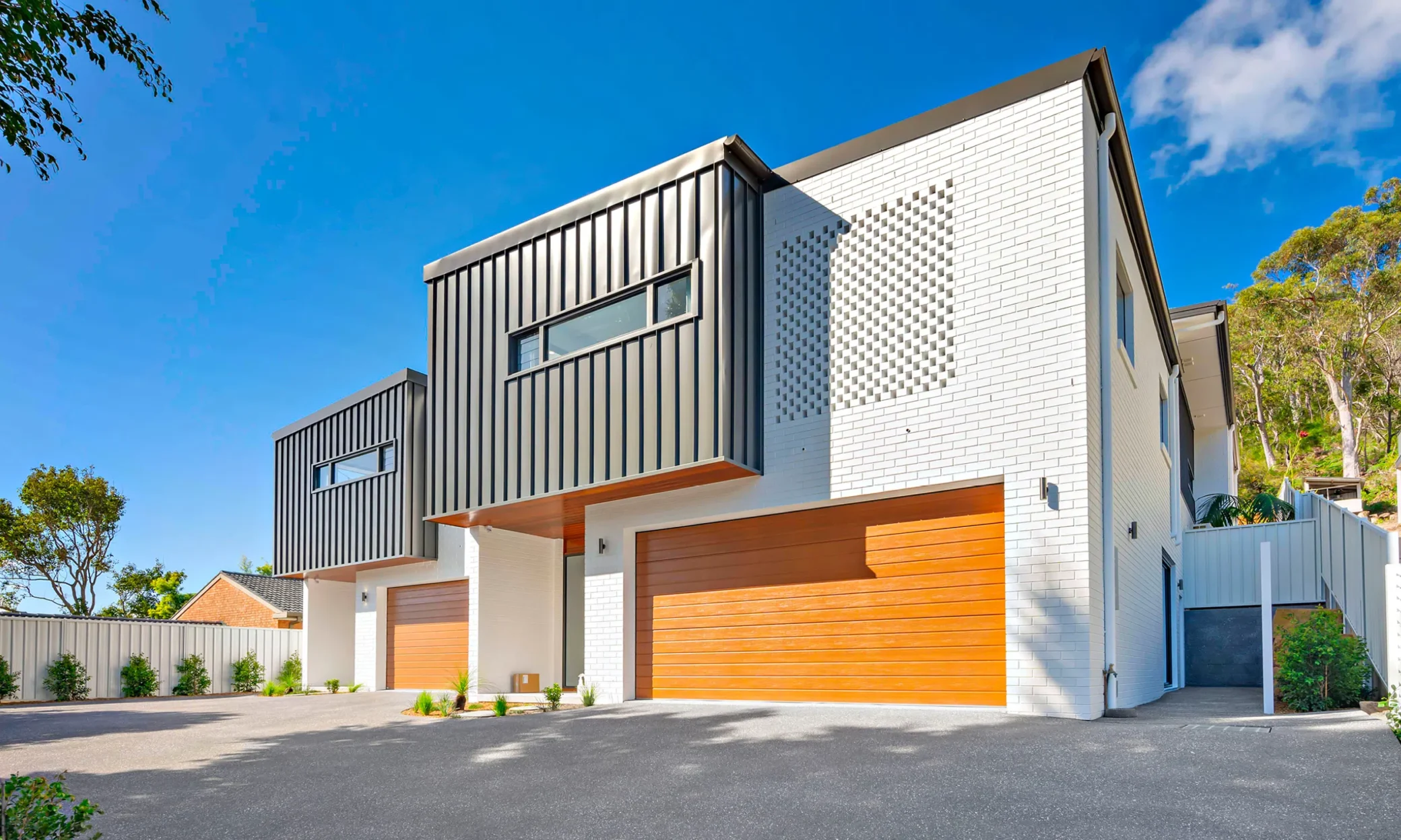 Modern two-story house in Nelson Bay with a geometric design, featuring a combination of white walls and dark gray metallic panels. It has wooden garage doors and is surrounded by green foliage under a clear blue sky