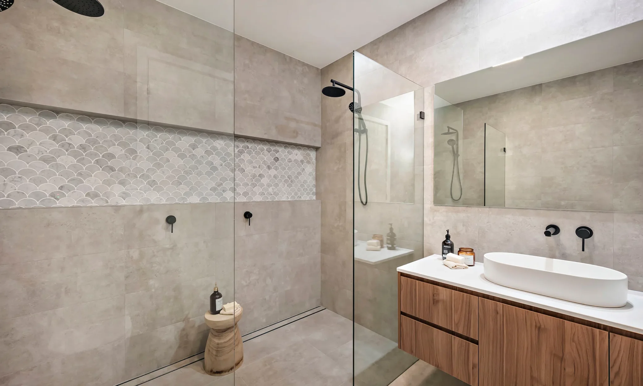 Modern bathroom in Newcastle featuring beige tiles, wooden vanity with two sinks, wall-mounted faucets, large mirror, and a walk-in shower with glass door and dual shower heads.