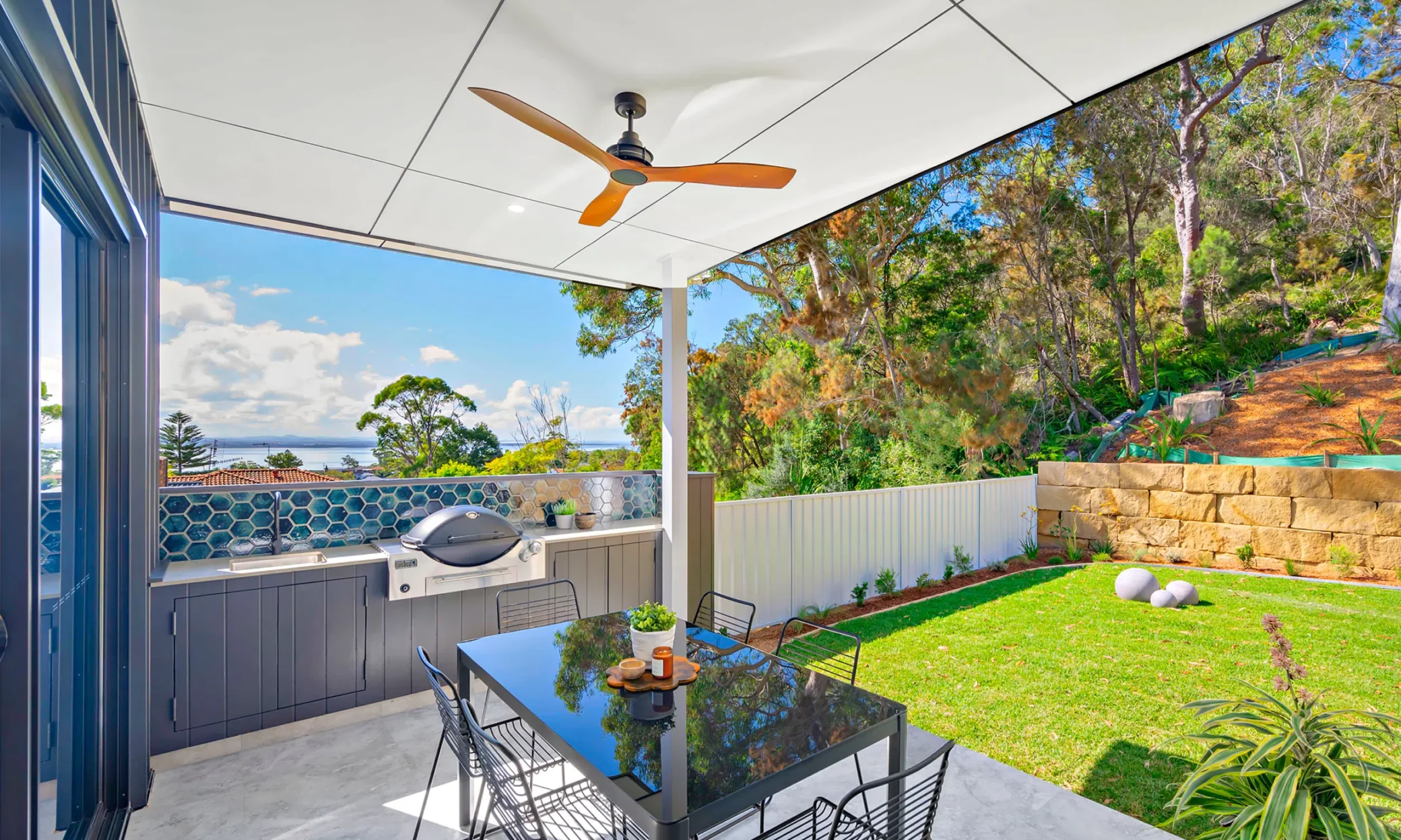 A modern outdoor patio in Port Stephens with a ceiling fan, dining area, barbecue grill, surrounded by lush greenery and a partial ocean view in the background, featuring vibrant blue accents on the furniture and