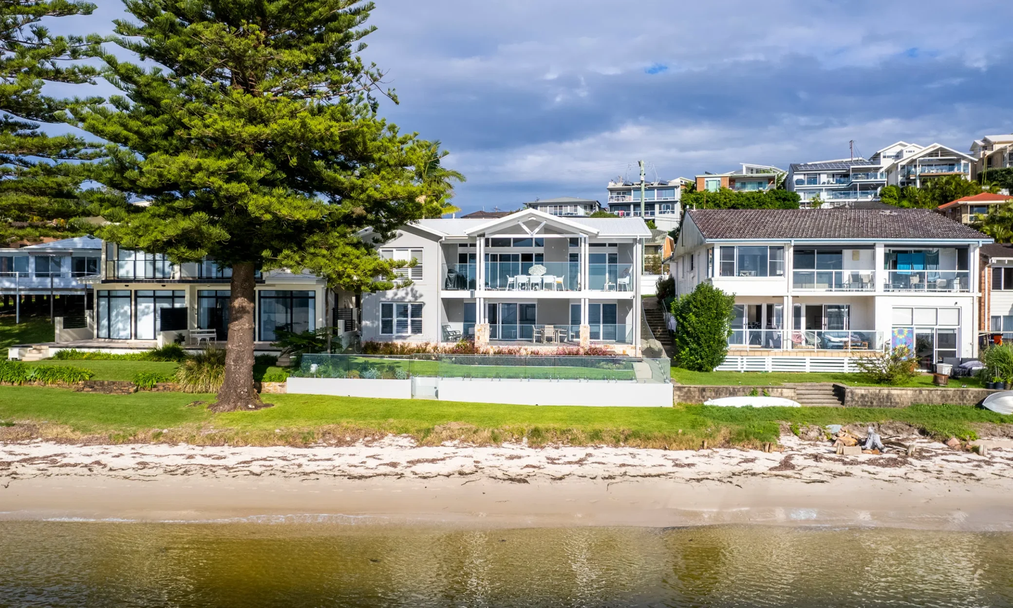 Luxury beachfront houses in Nelson Bay with large glass balconies overlooking a sandy shore, flanked by a lush green tree on the left. The houses feature contemporary architecture and are situated under a partly
