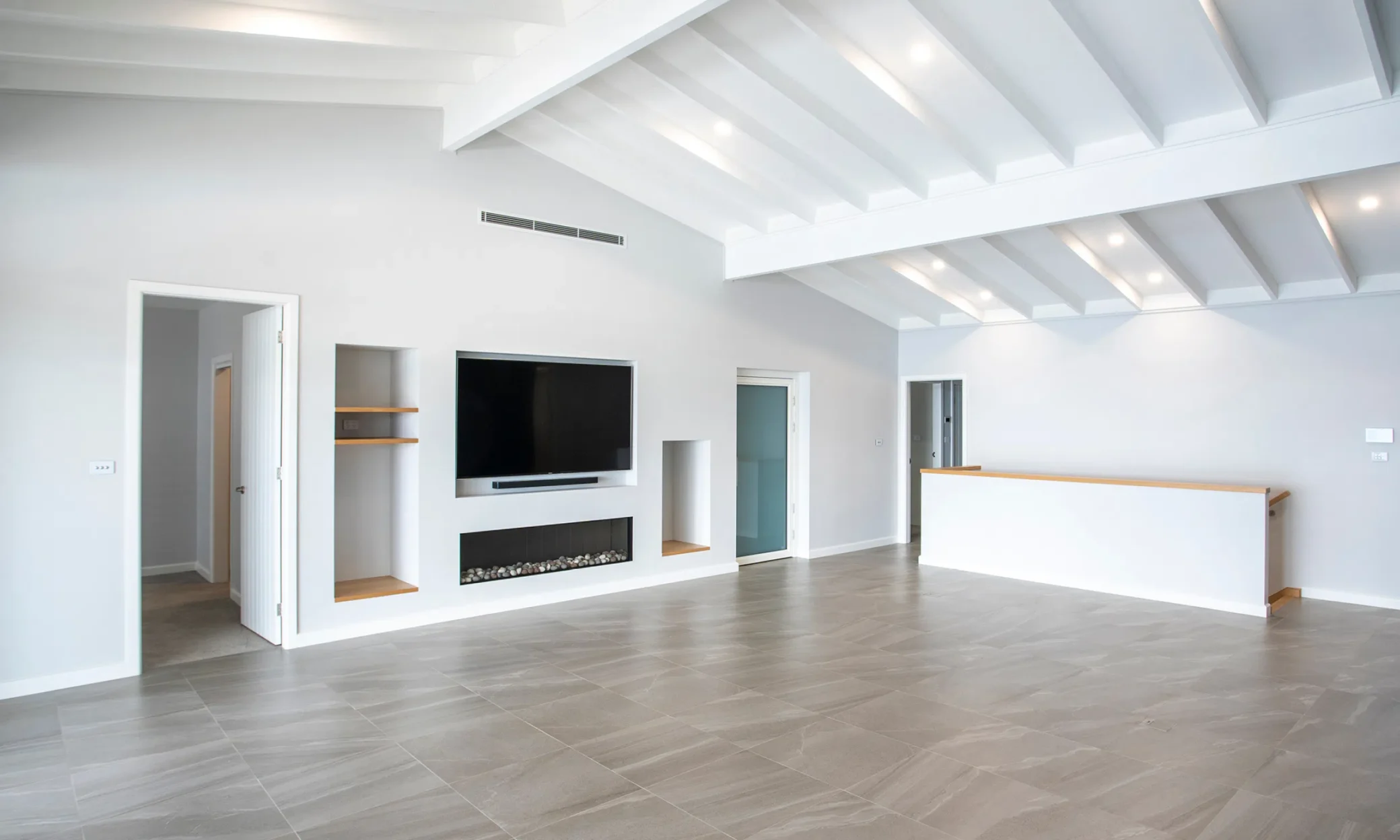 A modern, spacious living room with gray tiled flooring, white walls, and a high ceiling with exposed white beams. There is a built-in fireplace under a wall-mounted TV, and to the right,