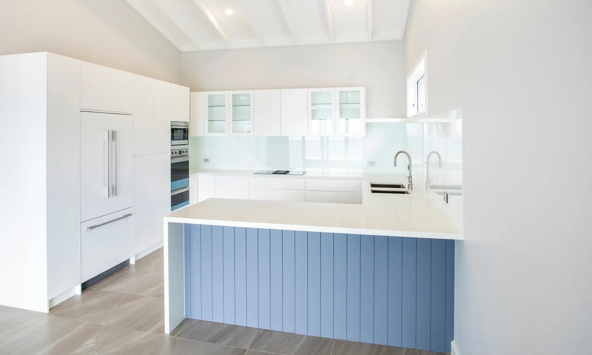 Modern kitchen with white and light blue cabinetry, stainless steel appliances, and gray tile floors. There's a central island with a sink under bright natural light, perfect for custom built projects in