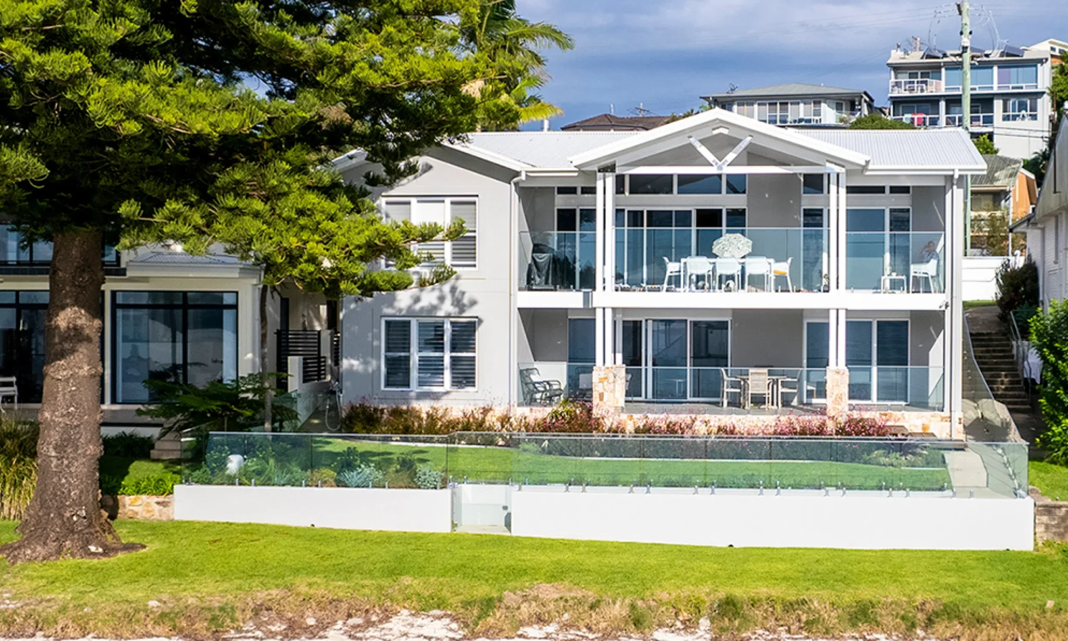 Modern two-story beachfront house in Port Stephens with large windows, a balcony, and a glass fence. There's a prominent pine tree to the left and grass in the foreground.