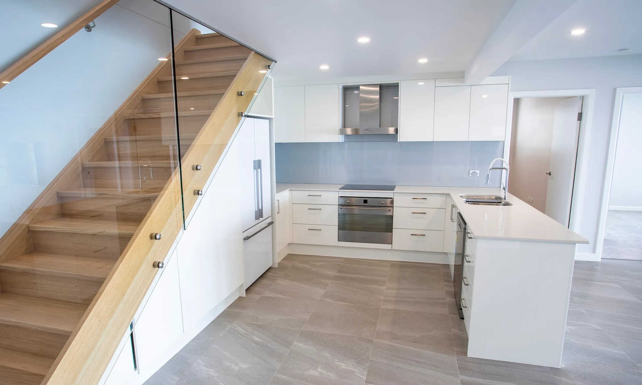 A modern kitchen with white cabinets and a central island next to a wooden staircase with glass railings, featuring built-in appliances and blue backsplash. Light gray tiled flooring extends throughout the Newcastle space.