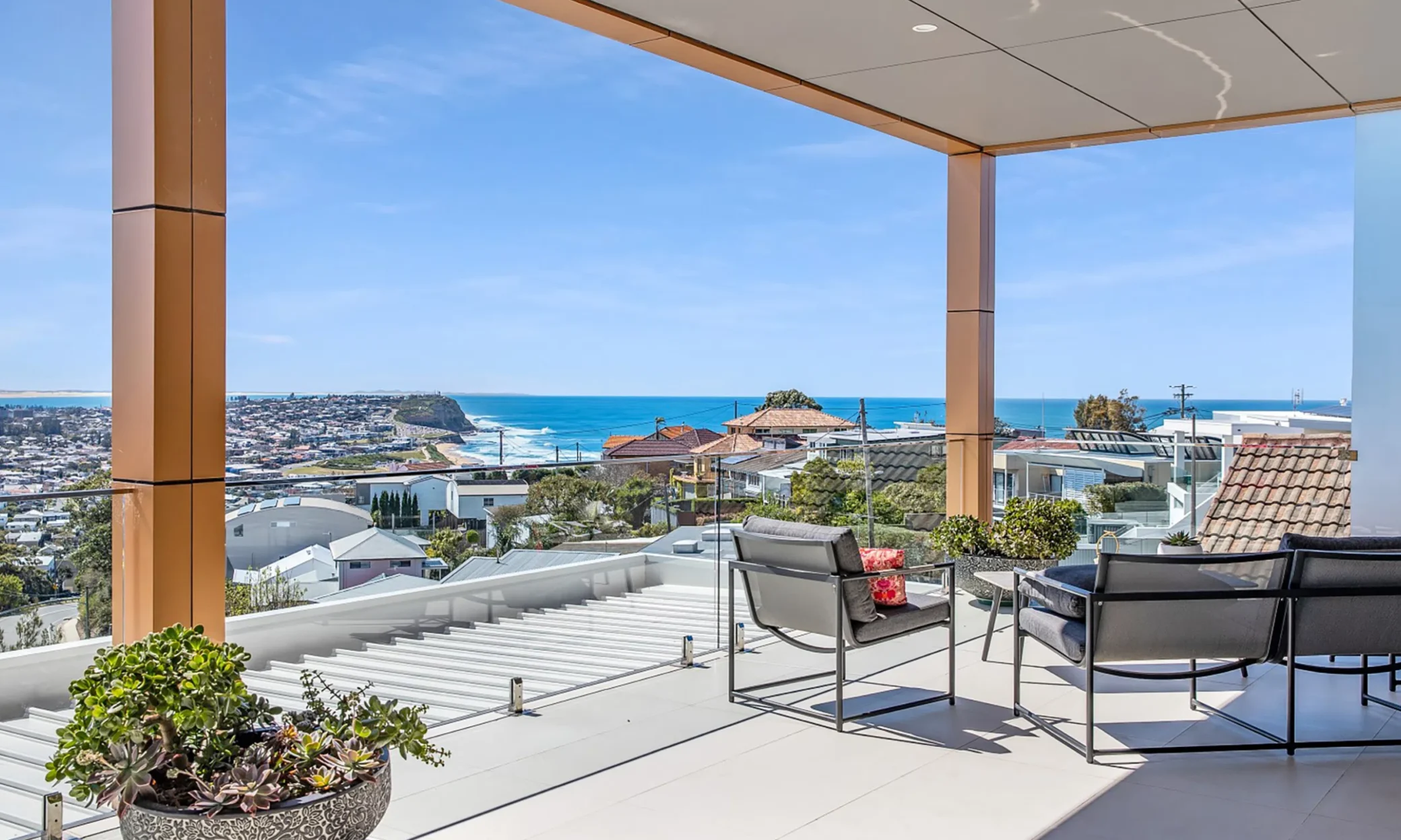 A modern terrace with a glass balustrade offers a stunning panoramic view of Newcastle and the ocean. The space is furnished with a dark table, chairs, and decorative plants, under a clear blue sky