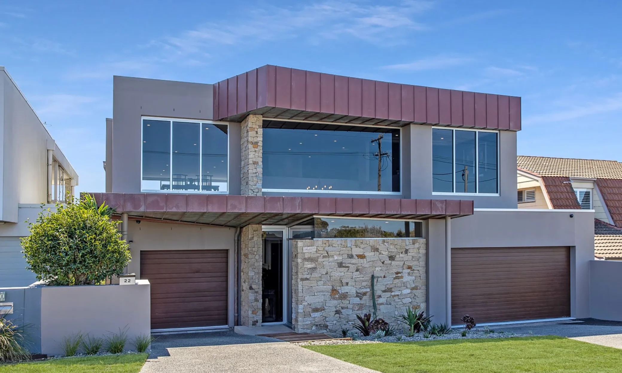 Modern two-story house with a flat roof and large glass windows, located in Port Stephens. Features a reddish-brown upper facade, double garage doors, and a stone-clad entrance. Landsc