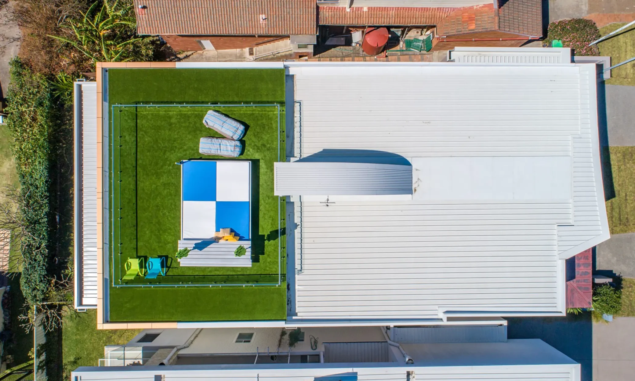 Aerial view of a residential property in Newcastle with a white, flat-roofed house featuring a rooftop mini soccer field surrounded by high walls, adjacent to other homes.