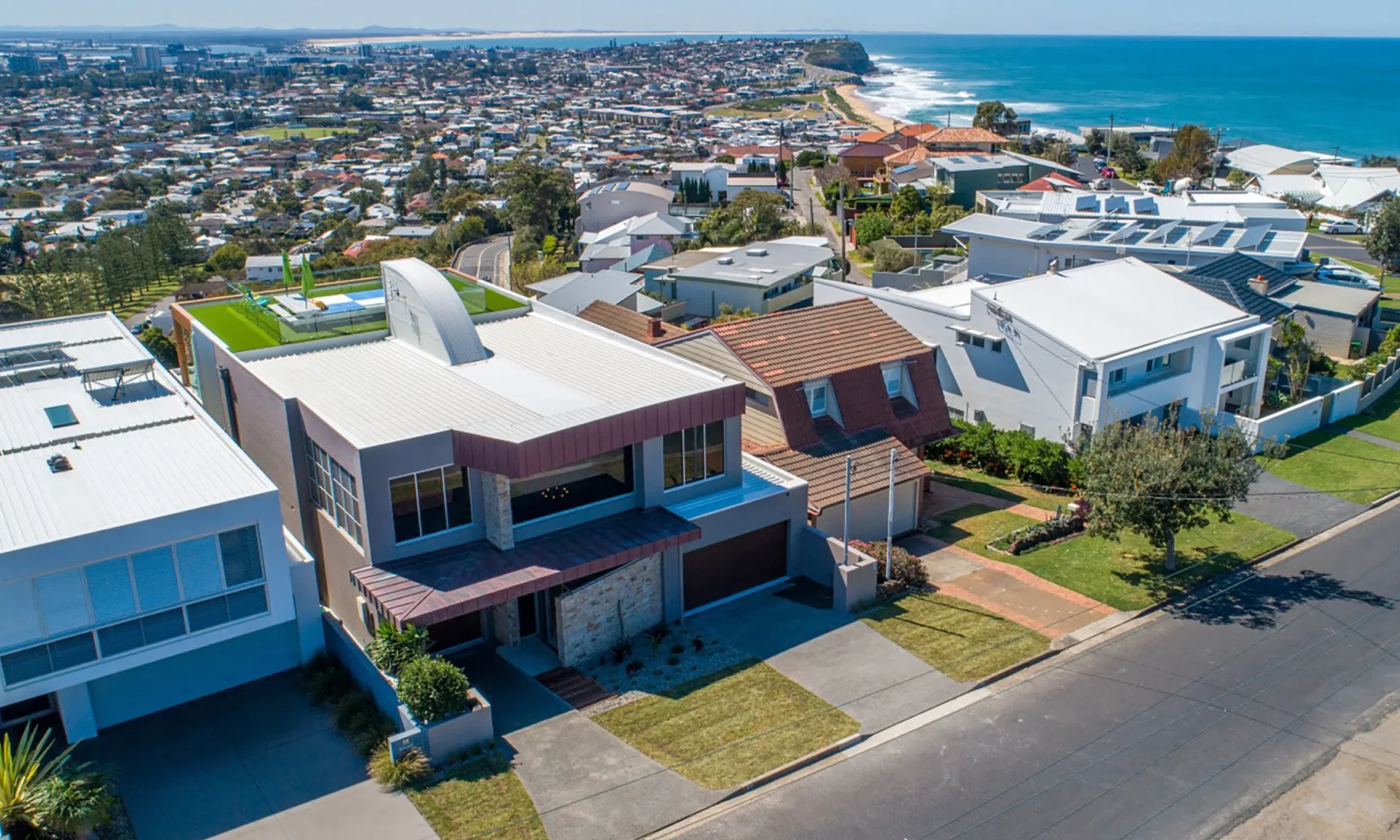 Aerial view of a coastal neighborhood in Port Stephens featuring modern houses with varying designs, predominantly in neutral colors, situated close to each other along a street, overlooking a scenic ocean in the background.