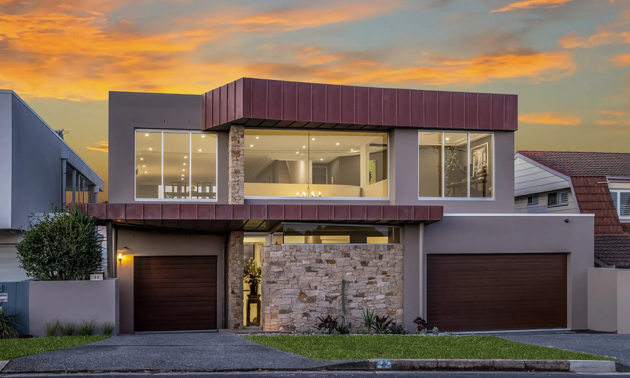 Modern two-story house featuring a striking upper facade with large glass windows and a balcony, grounded by stone-clad and wooden garage doors, set against a dramatic sunset sky with orange and pink hues in Newcastle