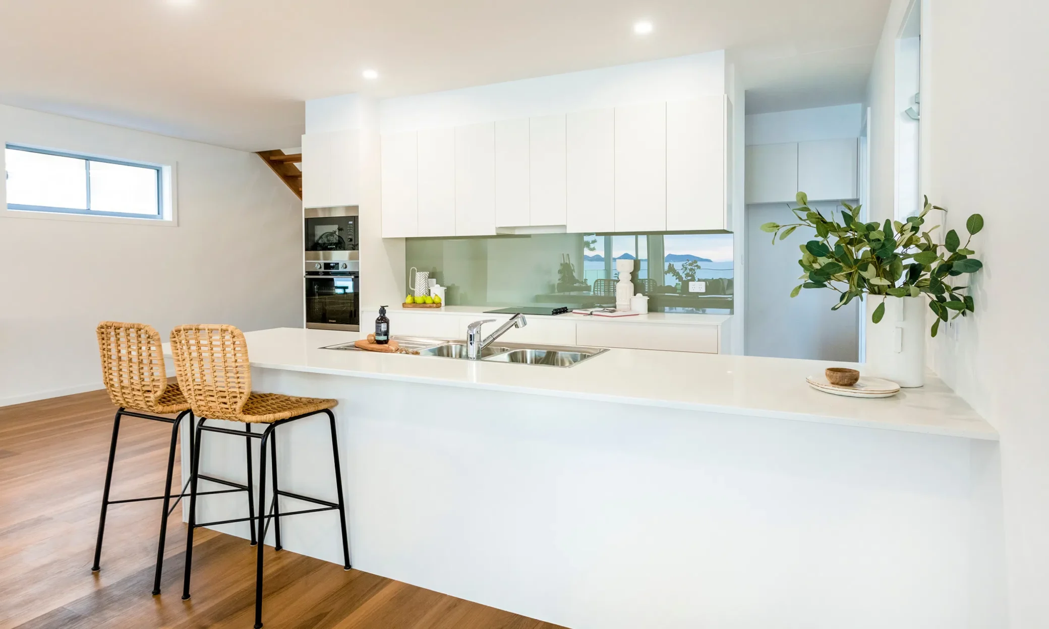 A modern kitchen featuring white cabinetry and countertops, with a central island and two wicker bar stools. Wooden flooring complements the room. A green potted plant and a reflective backsplash add to