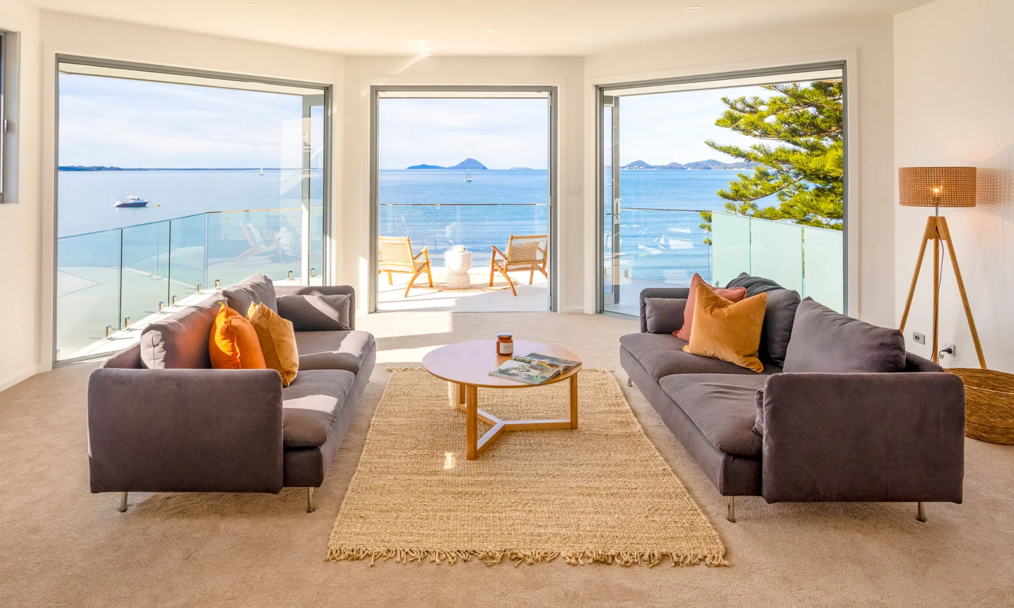 A bright, modern living room with two gray sofas facing each other over a beige rug, large glass doors open to a balcony overlooking Port Stephens with islands in the distance, and a clear blue sky.