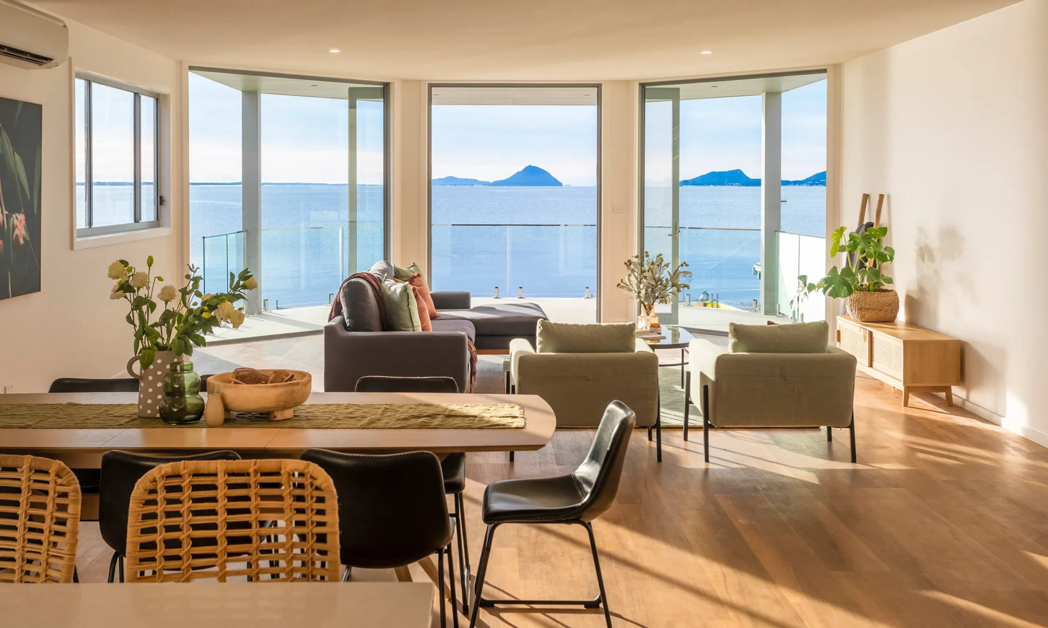 A modern living room in Nelson Bay with large windows offering a panoramic view of a calm sea and distant mountains. The room features a dining table, chairs, a sofa, armchairs, and p
