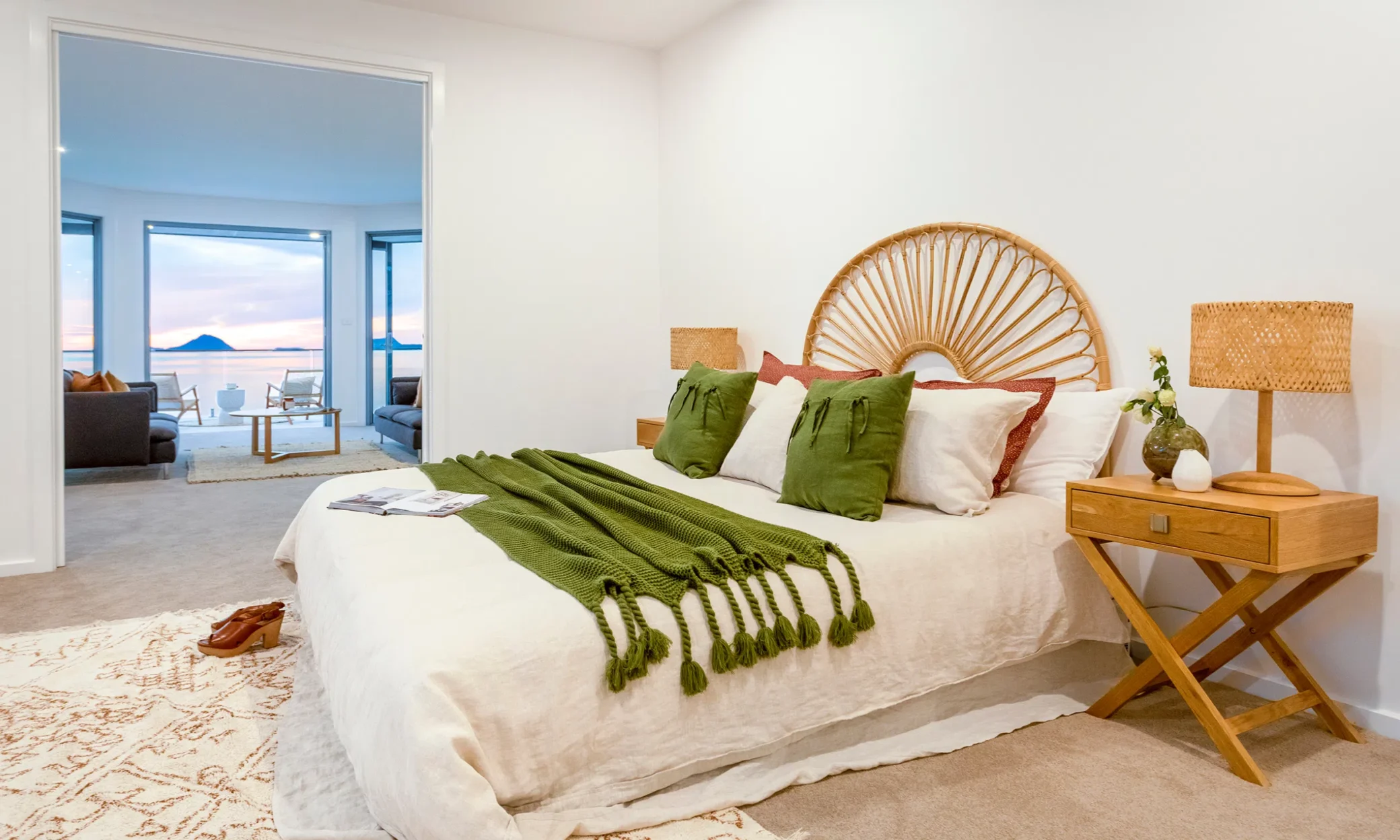 A serene bedroom in Nelson Bay with a large bed adorned with white and green cushions and a green throw, a rattan headboard, bedside table with lamp, a small seating area by a wide open