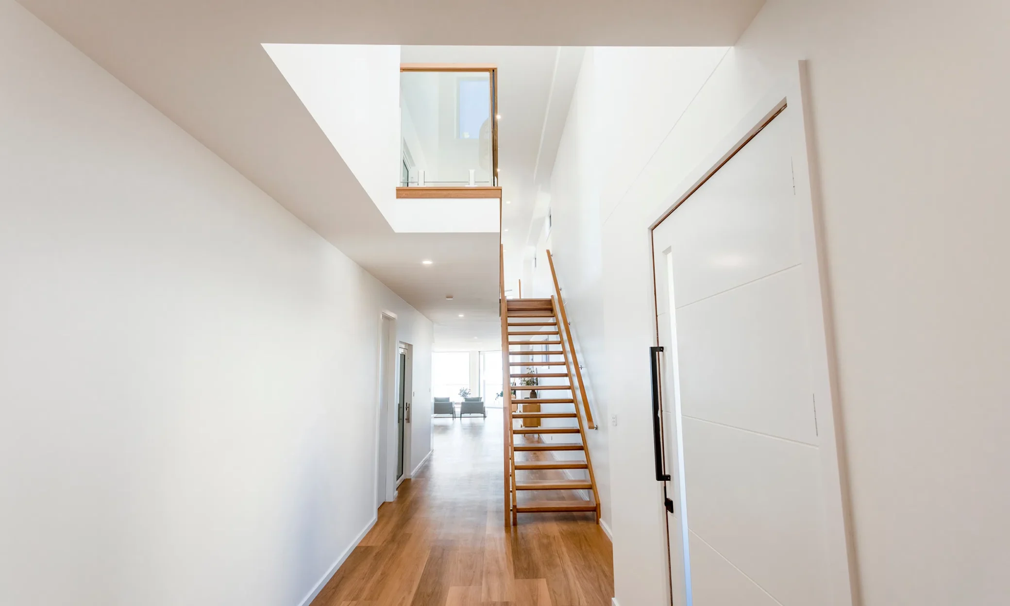 Modern hallway in a home in Newcastle with bright white walls and wooden flooring. It includes a skylight, a wooden staircase to the right leading up to an upper level, and a view into a