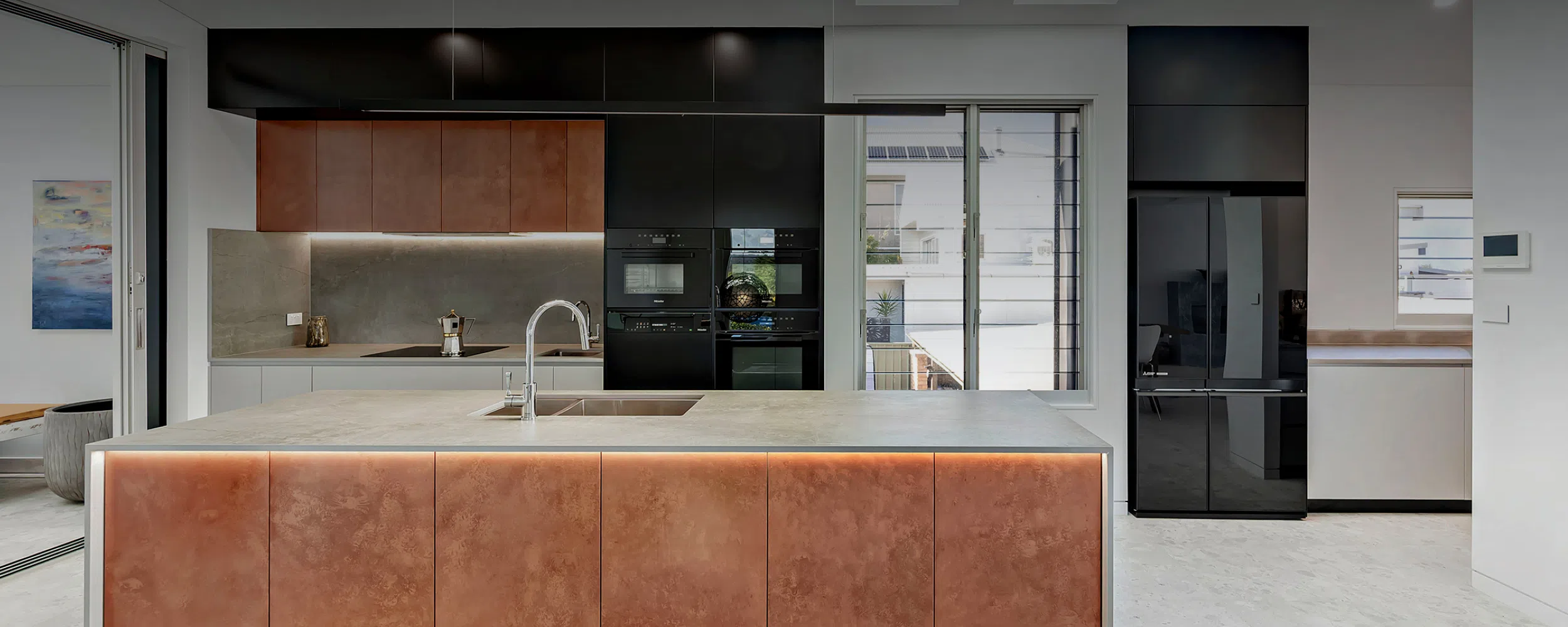 A modern kitchen featuring a large copper island, sleek black cabinetry, integrated appliances, and a light gray tiled floor. Natural light from a window illuminates the space in this custom-built project in Nelson