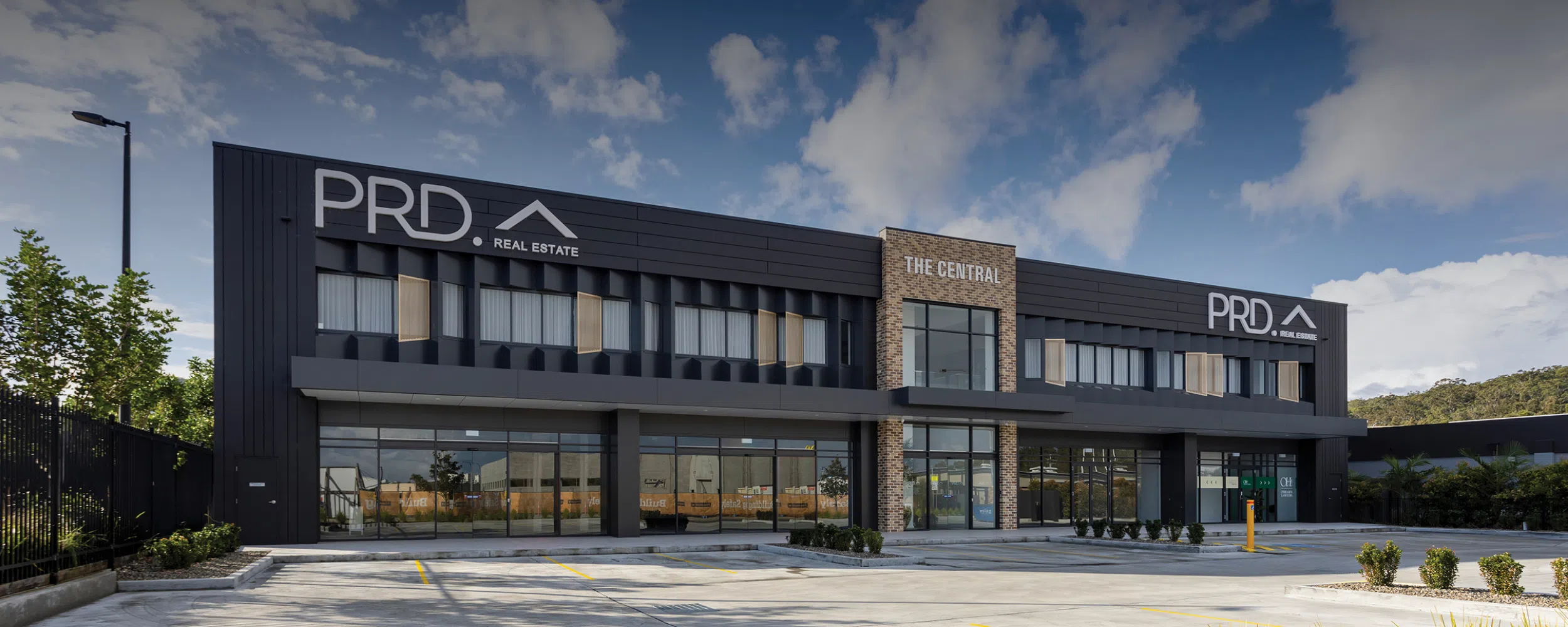 Modern two-story commercial building with signage for "prd real estate" and "the central." The structure, a custom-built project in Newcastle, features sleek black paneling with glass windows. An empty parking