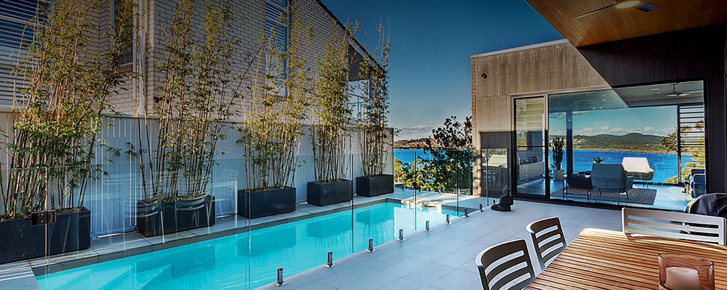 A modern terrace featuring a clear blue infinity pool bordered by glass barriers and potted plants. This custom-built project includes outdoor seating areas and opens to a panoramic ocean view, complemented by a clear sky