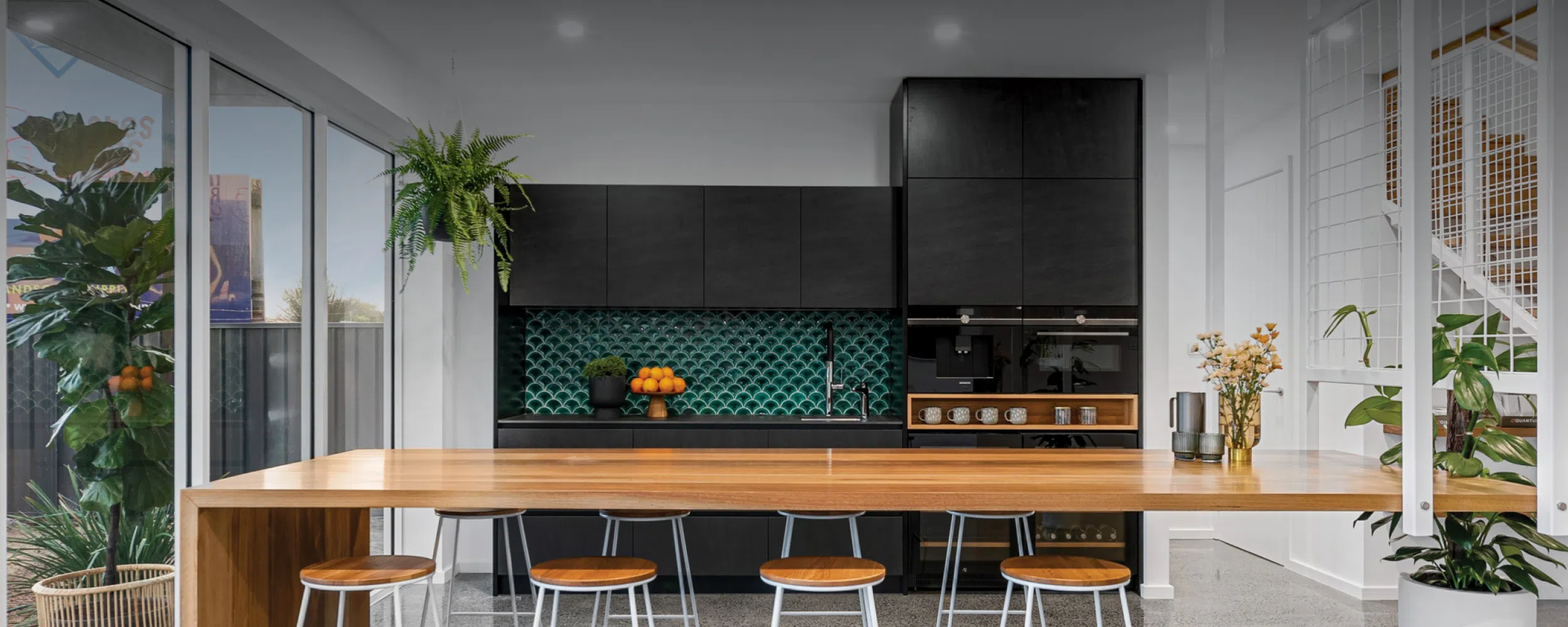 A modern kitchen featuring black cabinets and a vivid green tile backsplash, with a large wooden dining table, high stools, and decorative plants. Bright and airy due to large windows and soft natural lighting.