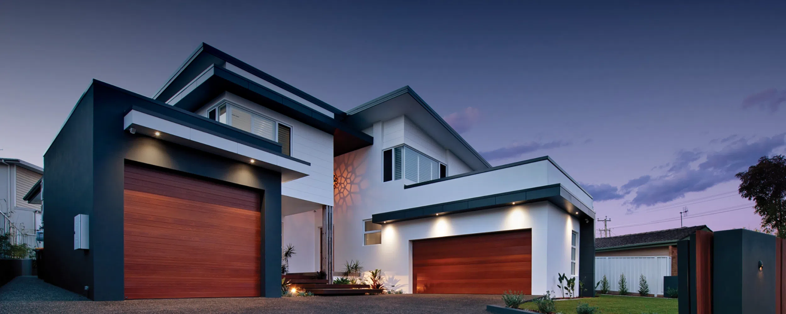 A modern two-story house at twilight in Nelson Bay with illuminated exterior lights, featuring a mix of dark and light gray walls, two large redwood garage doors, and a neat front yard.