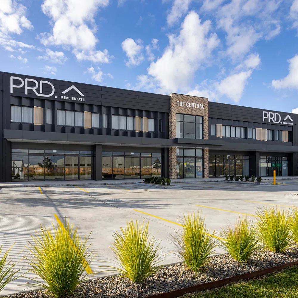 Modern two-story commercial building with the signage "prd real estate" and "the central." features a dark gray facade, large glass windows, landscaped front with green plants, and an empty parking lot