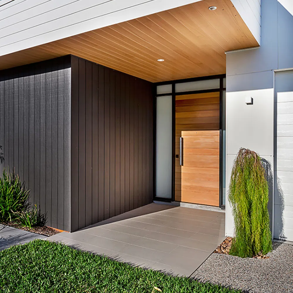 Modern house entryway in Nelson Bay featuring a combination of dark vertical siding and white smooth walls, with a wooden door accentuated by glass panels. The porch has neat concrete flooring and is flanked by