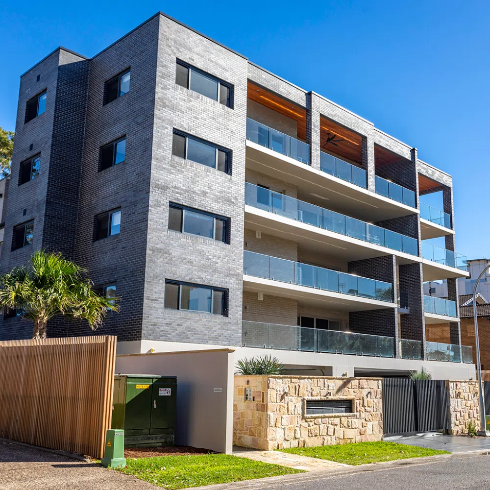 Modern multi-story residential building with a mix of gray brick and warm wooden panels. Features balconies, large windows, and protected parking at street level. A sunny day enhances the clean, angular architecture of