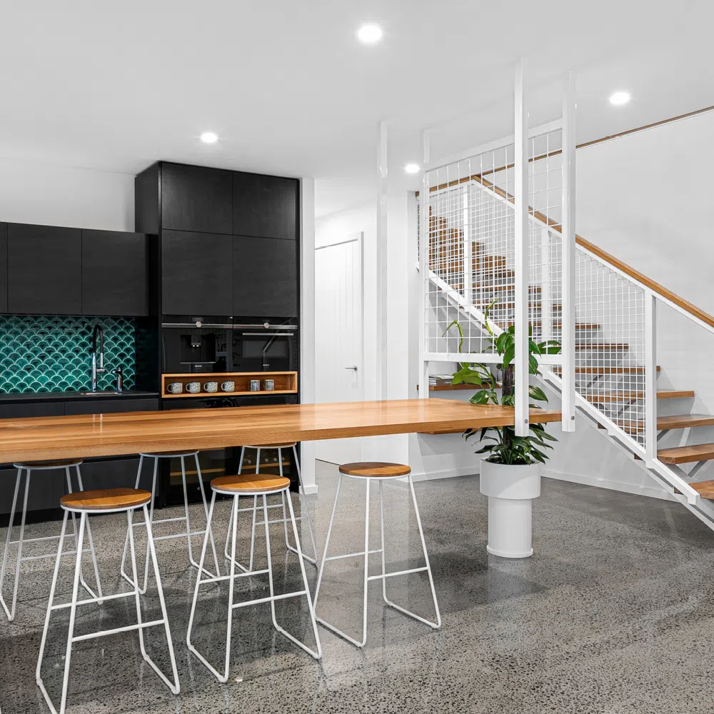 Modern kitchen with black cabinetry and a teal tile backsplash in Newcastle. A wooden bar counter with white stools extends towards a staircase with white railing and wooden steps. The floor is speckled gray