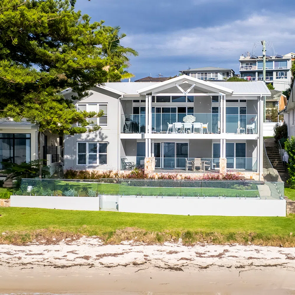 Modern beachfront house in Nelson Bay with large windows, multiple balconies, and a clear fence. The house is flanked by lush green trees and has direct beach access, with the ocean visible in