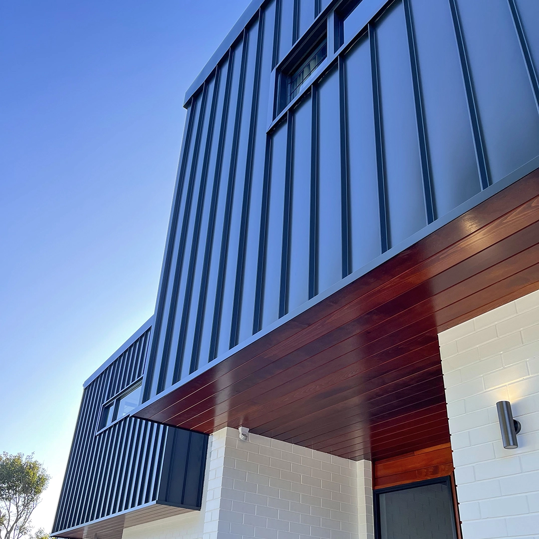 A modern building facade with vertical metal siding, part of the structure features red wooden panels; set against a clear blue sky. Bright sunlight highlights the sleek architectural details.