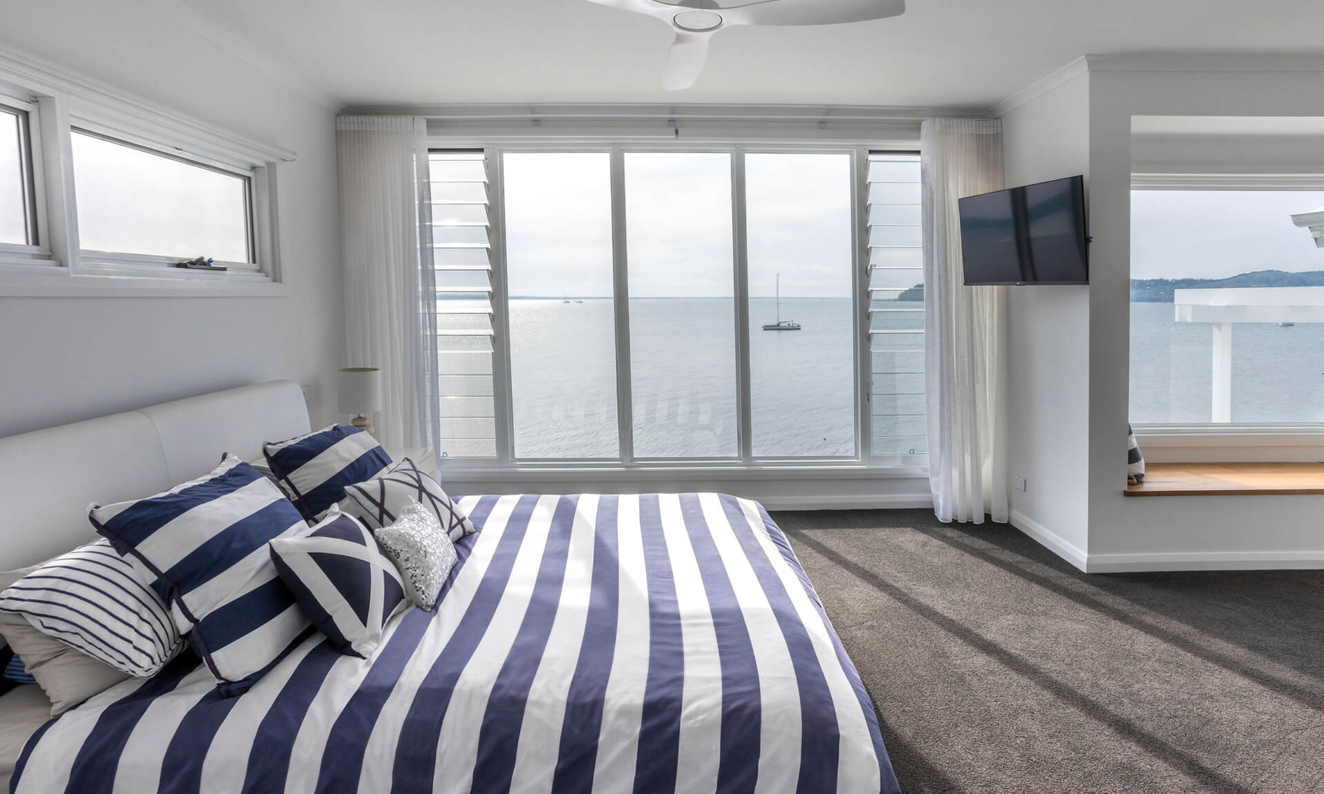 A modern bedroom with blue and white striped bedding, large windows offering a panoramic view of the sea with boats, white walls, a ceiling fan, and a mounted TV on the right.