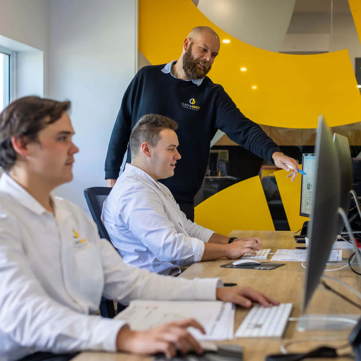 Three men in a modern office setting in Nelson Bay, with one standing and pointing at a computer screen, and two others, seated, looking on attentively. They wear smart casual attire, and the